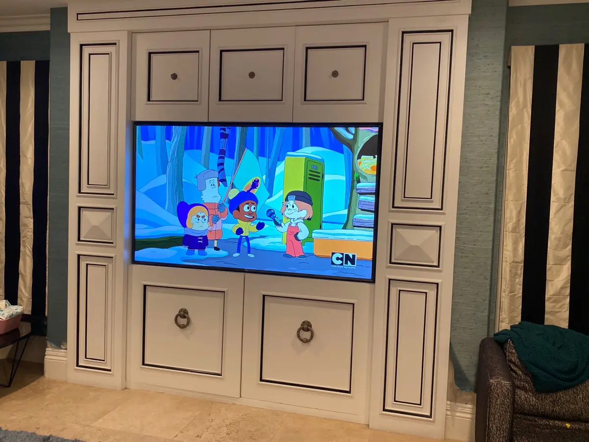 A flat screen TV installation is showcased on a white built-in entertainment center, displaying a cartoon scene with four characters. The room features tiled flooring and drapery gracefully hanging on each side of the entertainment center.