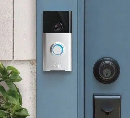 A silver and black Ring video doorbell, a key feature of Smart Home Solutions, is mounted next to a blue door with a silver handle and lock. A green plant is visible on the left side.