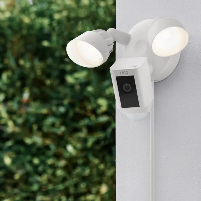 A Ring floodlight security camera, a key component of Smart Home Solutions, is mounted on an exterior wall. The camera has two LED lights and faces outward towards a leafy background.