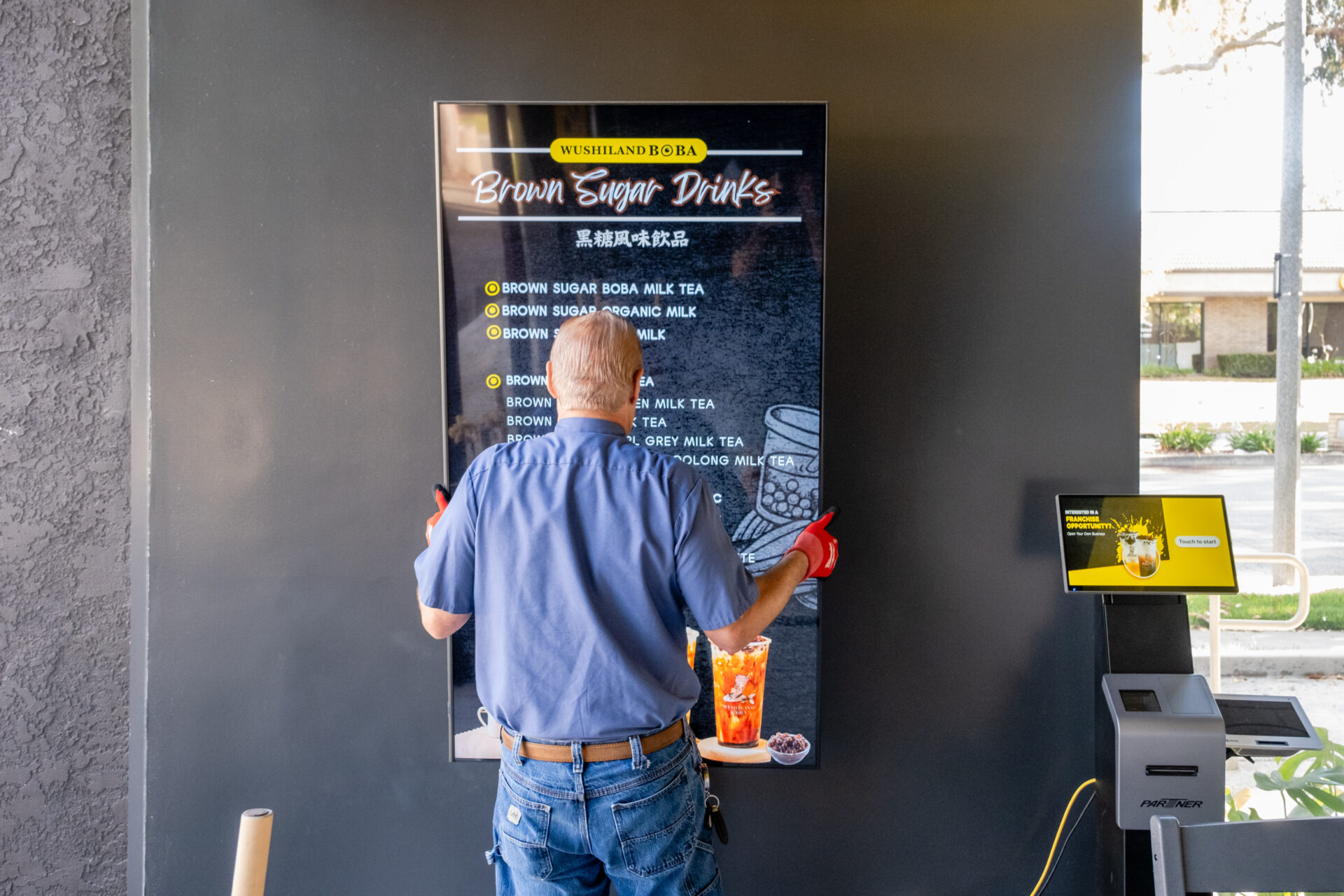 A person in a blue shirt adjusts a digital menu display for brown sugar drinks at a shop. The setup, reminiscent of home automation services, appears to be in progress with the person facing away from the camera.